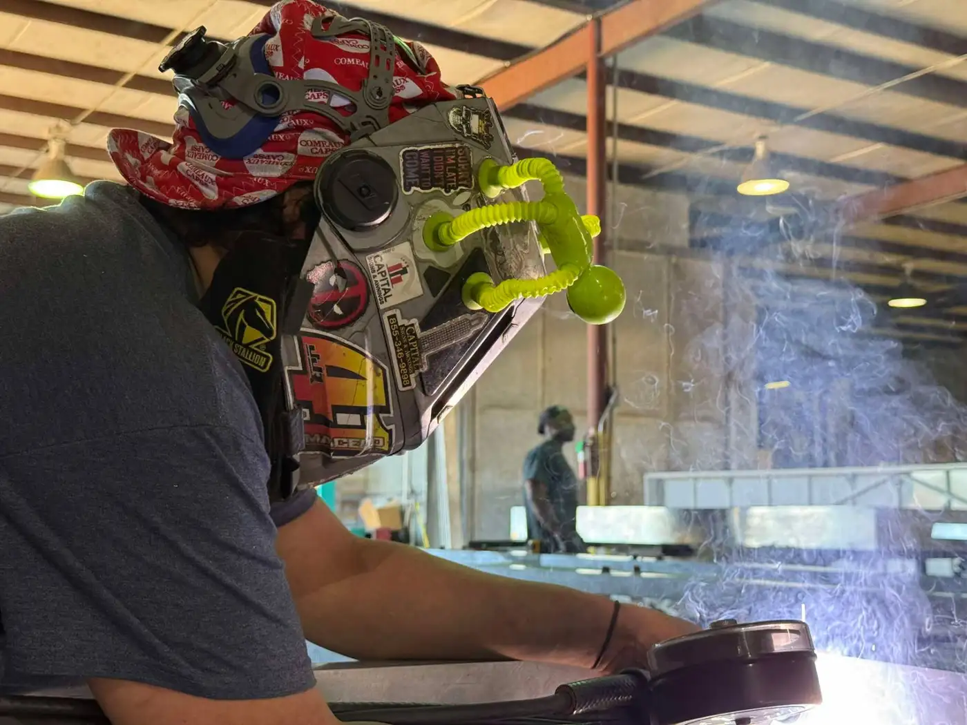 Welder in a hood MIG welding a large sign frame with an arc flash and smoke inside the fabrication shop